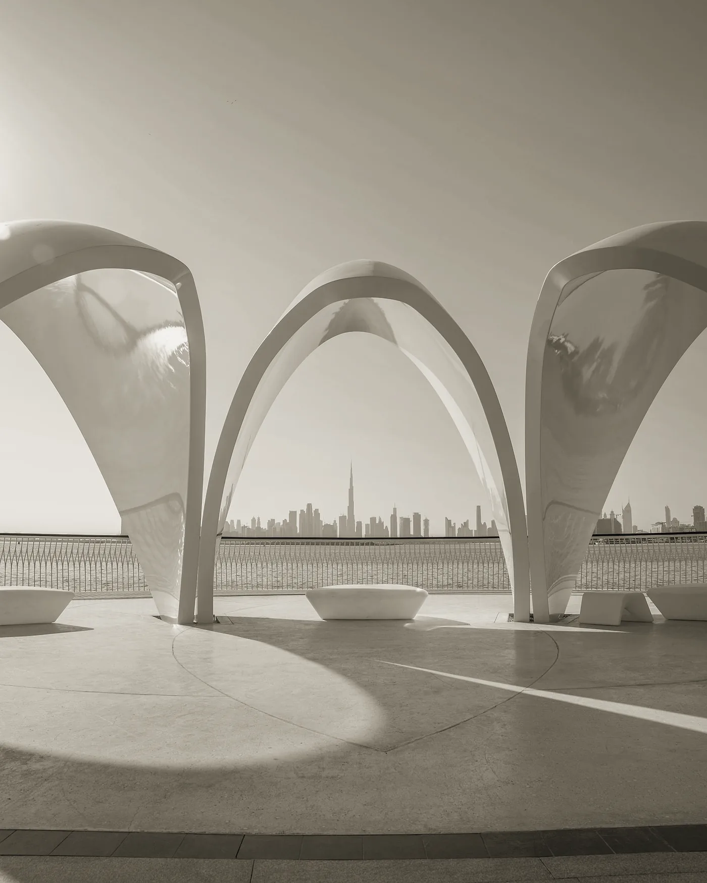 Arches blanches d’un pavillon en bord de mer à Dubaï, la ligne d’horizon de la ville au loin, sous un soleil éclatant.